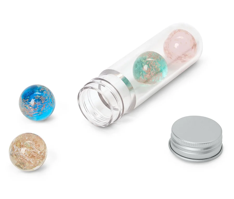 Quatre sphères pailletées bleues, turquoise, roses et beiges, dont deux à l'intérieur d'un tube en plastique transparent ouvert et deux à l'extérieur. Un capuchon argenté est posé à côté du tube.