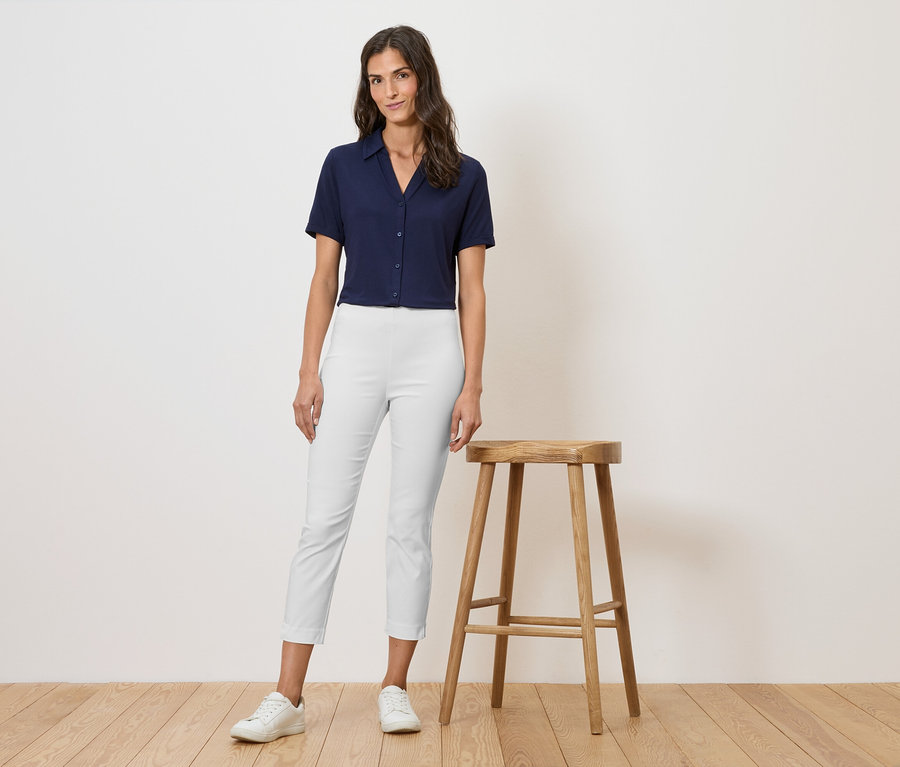 Femme en chemise bleu marine et pantalon blanc debout à côté d'un tabouret en bois.