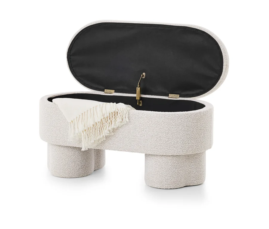 Pouf de rangement ovale en tissu bouclé crème, avec son couvercle ouvert révélant un intérieur sombre. Une couverture crème à franges est drapée sur le bord du pouf, qui repose sur quatre pieds épais et arrondis.