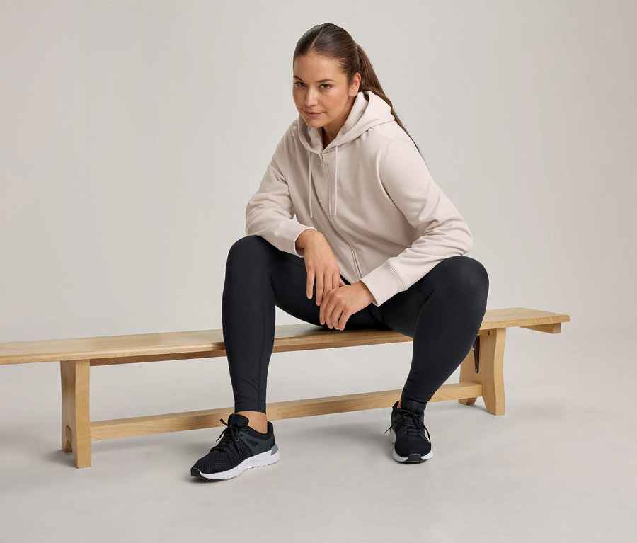 Femme assise sur un banc en bois, portant un sweat à capuche zippé beige clair, un legging noir et des chaussures de sport noires.