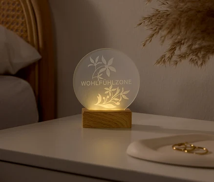 Lampe ronde lumineuse avec une base en bois sur une table de chevet blanche. La lampe présente des motifs floraux gravés et le texte « WOHLFÜHLZONE ». Une tête de lit en rotin et de l'herbe de la pampa sont visibles en arrière-plan.