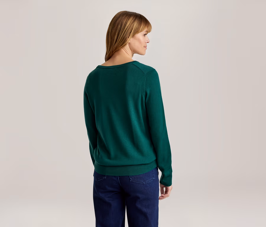 Femme vue de dos, légèrement tournée vers la droite, portant un pull vert à manches longues et un jean bleu foncé.