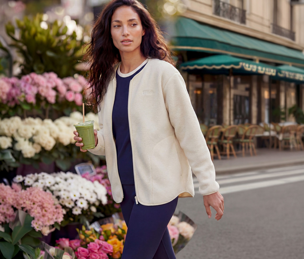 Une femme aux longs cheveux foncés marche dans une rue de la ville. Elle porte une veste polaire Fila crème sur un haut bleu marine et tient un smoothie vert. À l'arrière-plan, on voit un étal de fleurs et un café avec un auvent vert.