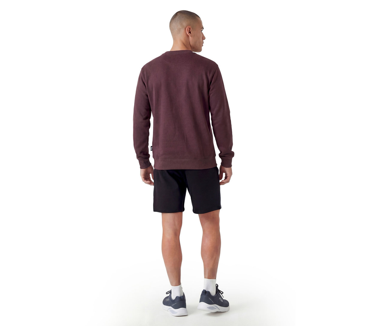 Homme debout de dos, portant un sweat-shirt bordeaux à col rond, un short noir, des chaussettes blanches et des chaussures de sport foncées.