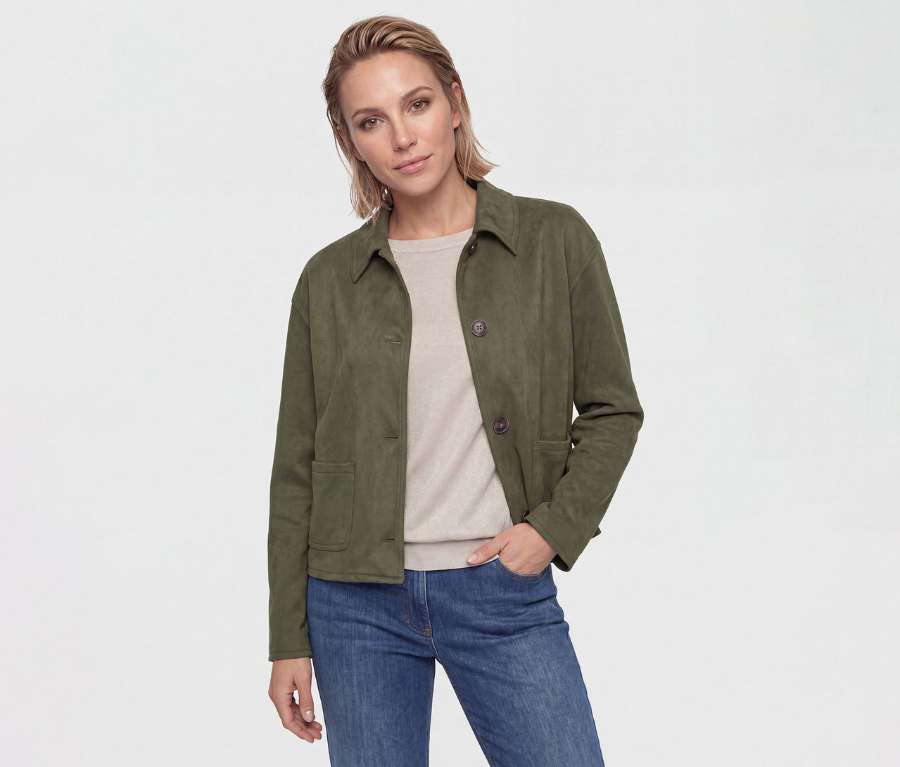 Une femme aux cheveux courts et châtains clairs porte une veste vert foncé, un haut beige et un jean bleu. Sa main est dans sa poche.