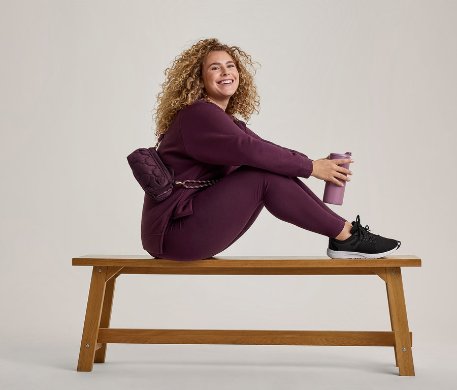 Une femme souriante aux cheveux bouclés est assise sur un banc en bois, vêtue d'un survêtement prune, d'un sac banane assorti et de baskets noires, tenant une bouteille d'eau violette.