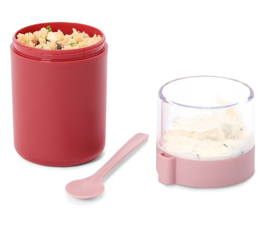 Récipient alimentaire rouge ouvert rempli de salade de céréales, un plus petit récipient rose et transparent avec une sauce crémeuse et une cuillère rose sur fond blanc.
