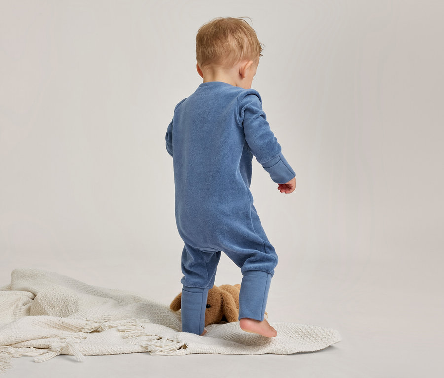 Un tout-petit, vu de dos, se tient sur une couverture blanche texturée, vêtu d'une combinaison bleue. Un animal en peluche marron est partiellement visible entre ses jambes.