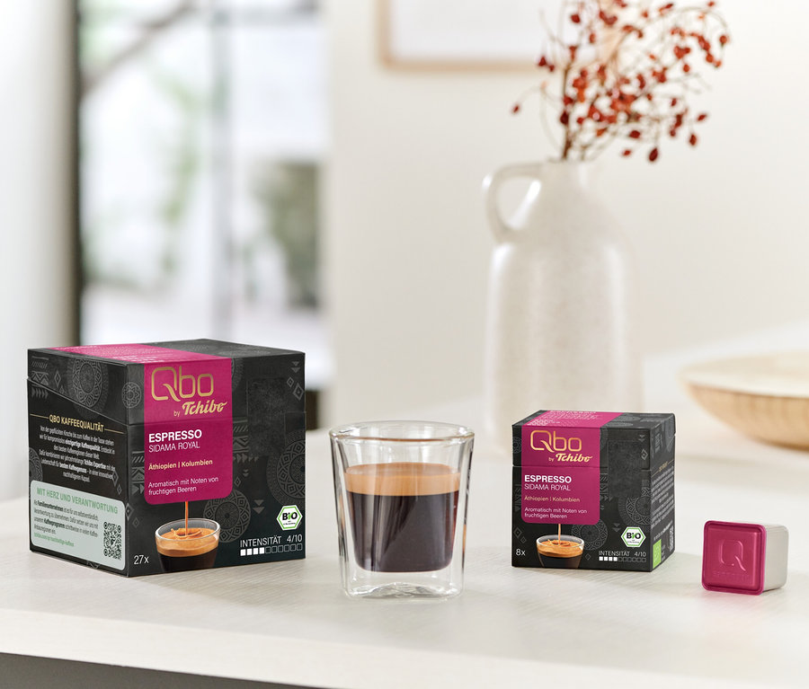 Deux boîtes de capsules Qbo Espresso SIDAMA ROYAL noires et roses (27 et 8 capsules), un verre d'espresso et une seule capsule rose et blanche sur une table blanche.