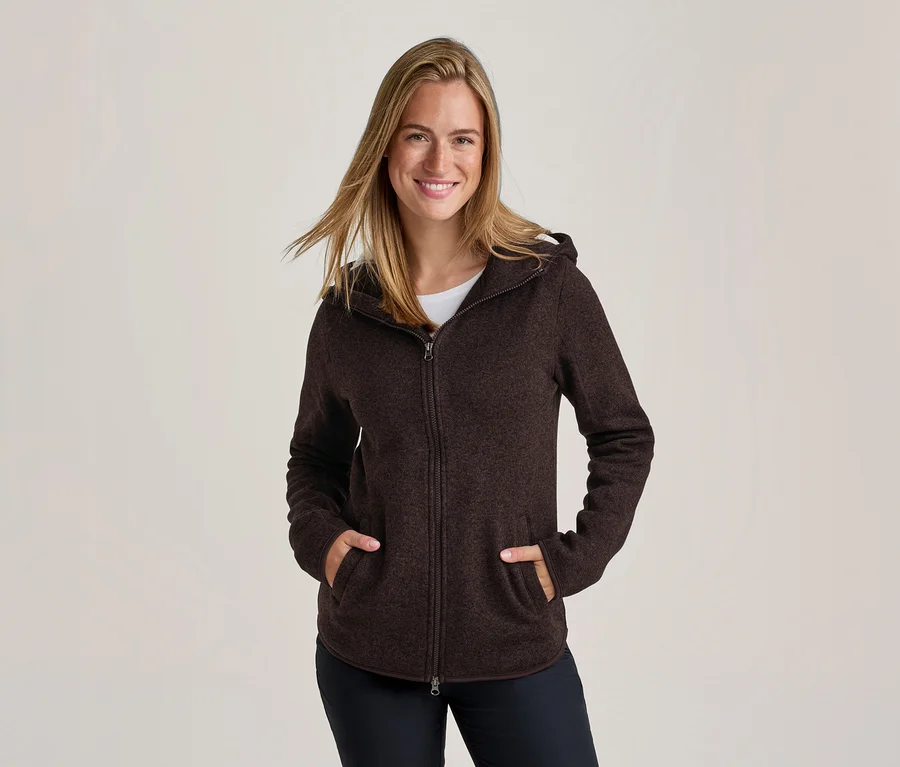 Femme souriante aux cheveux blonds portant un sweat à capuche zippé marron foncé, les mains dans les poches.