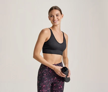 Femme souriante en brassière de sport noire et legging à motifs, tenant une gourde noire.