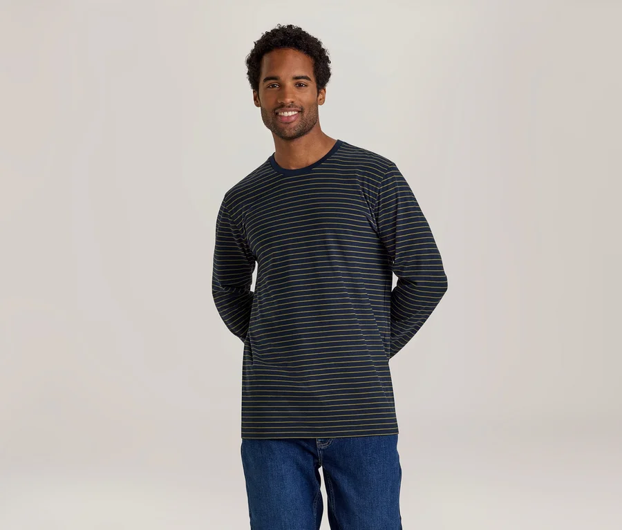 Un homme souriant aux cheveux bouclés foncés se tient devant un fond clair, portant un t-shirt à manches longues bleu marine à fines rayures beiges et un jean.