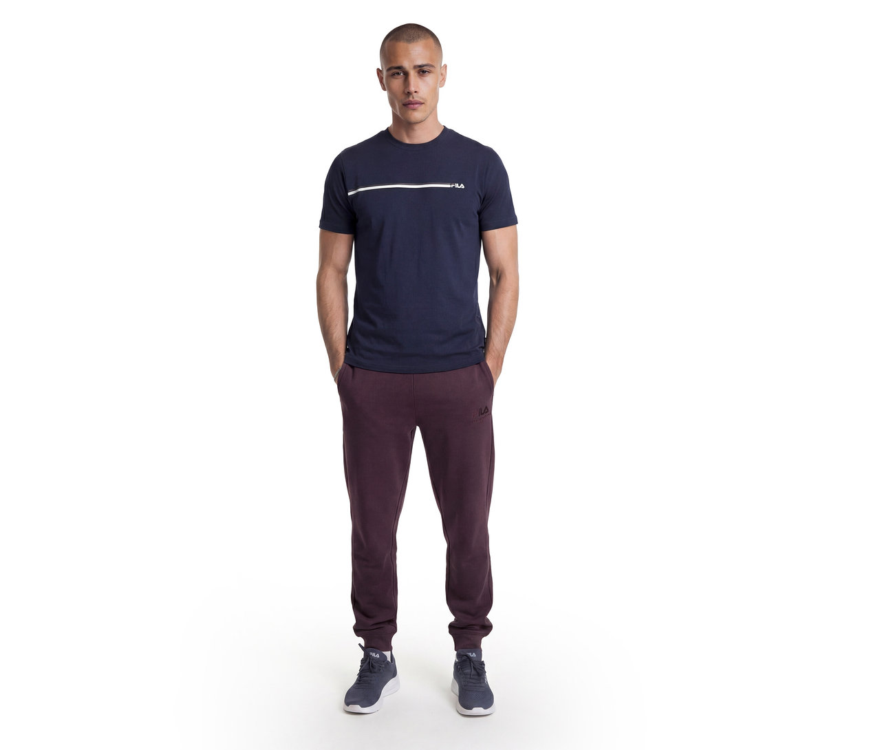 Homme debout, mains dans les poches, portant un t-shirt bleu marine avec une bande blanche et le logo Fila, un pantalon de jogging Fila bordeaux et des baskets bleu foncé.