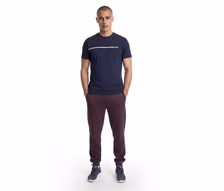 Homme debout, mains dans les poches, portant un t-shirt bleu marine avec une bande blanche et le logo Fila, un pantalon de jogging Fila bordeaux et des baskets bleu foncé.