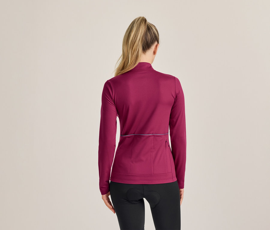Femme de dos, avec une queue de cheval blonde, portant un haut à manches longues magenta avec une bande réfléchissante et un cuissard noir.