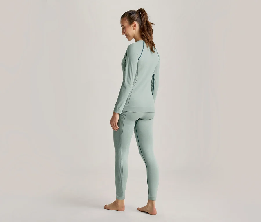 Femme vue de dos, légèrement de côté, portant un haut à manches longues vert clair et un legging assorti, les cheveux attachés en queue de cheval, pieds nus.