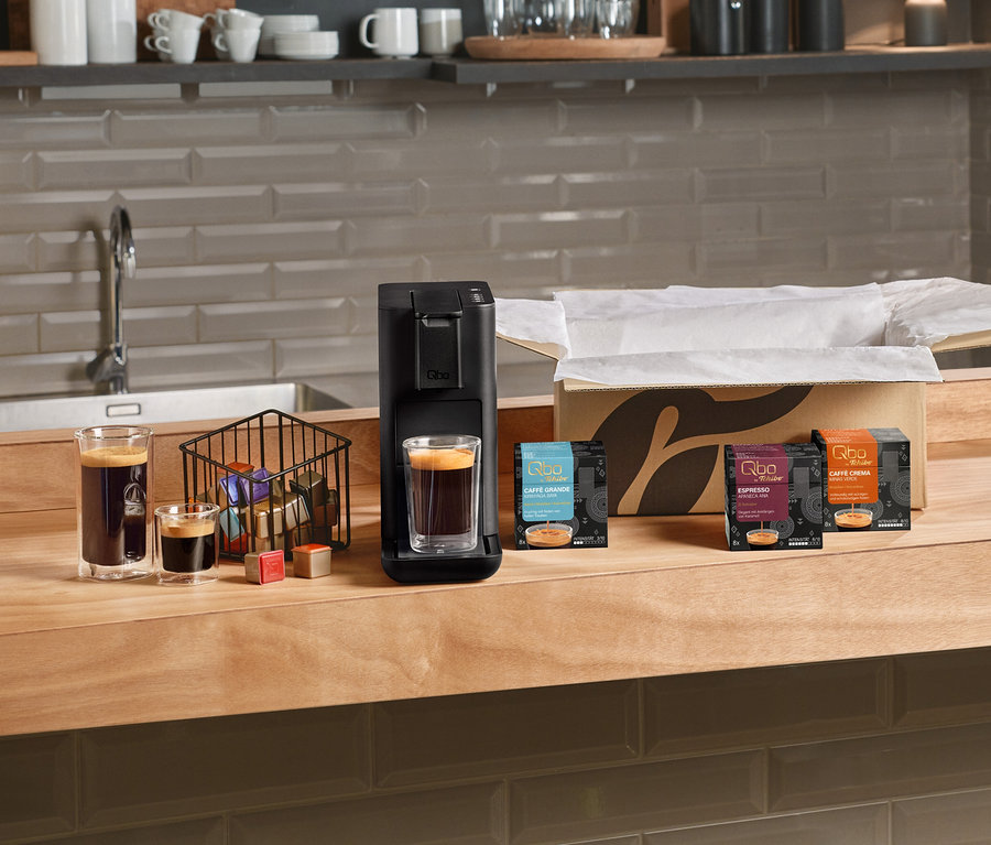 Une machine à café Qbo Iconic Dark Chrome noire avec un verre d'espresso, à côté d'un panier de capsules et de trois paquets de Qbo Caffè Grande Kirinyaga Baya, Qbo Espresso Apaneca Ana et Qbo Caffè Crema sur un comptoir en bois. Deux autres verres de café sont à gauche.