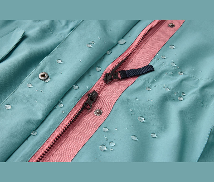 Gros plan d'un tissu imperméable bleu turquoise avec des gouttelettes d'eau, une fermeture éclair rose, des tirettes noires et des boutons-pression argentés.