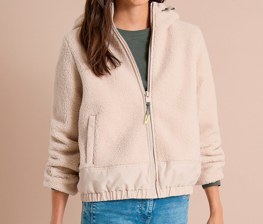 Femme souriante portant une veste CECIL beige zippée avec une partie supérieure moelleuse et une partie inférieure lisse, un haut vert et un jean bleu, tenant un article noir.