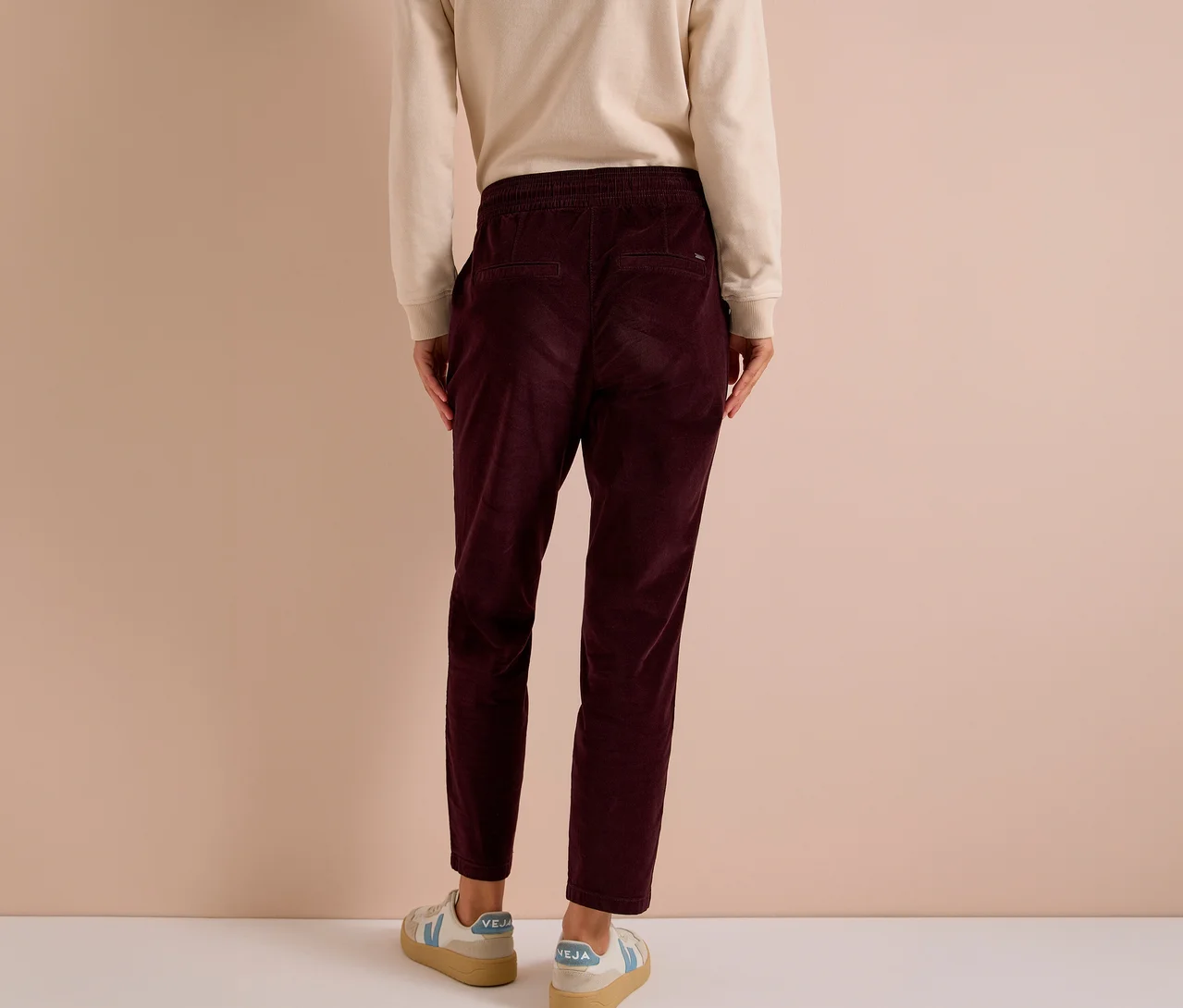Personne vue de dos portant un t-shirt clair à manches longues, un pantalon en velours côtelé CECIL rouge foncé avec une taille élastique et des baskets claires avec des accents bleus.