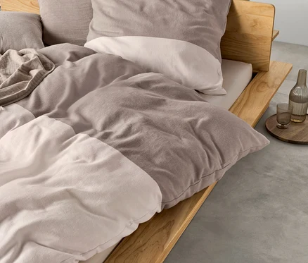 Un lit avec un cadre en bois clair, habillé d'une parure de lit en flanelle marron clair-taupe et d'une couverture plus claire. À droite, un plateau avec une bouteille et un verre.
