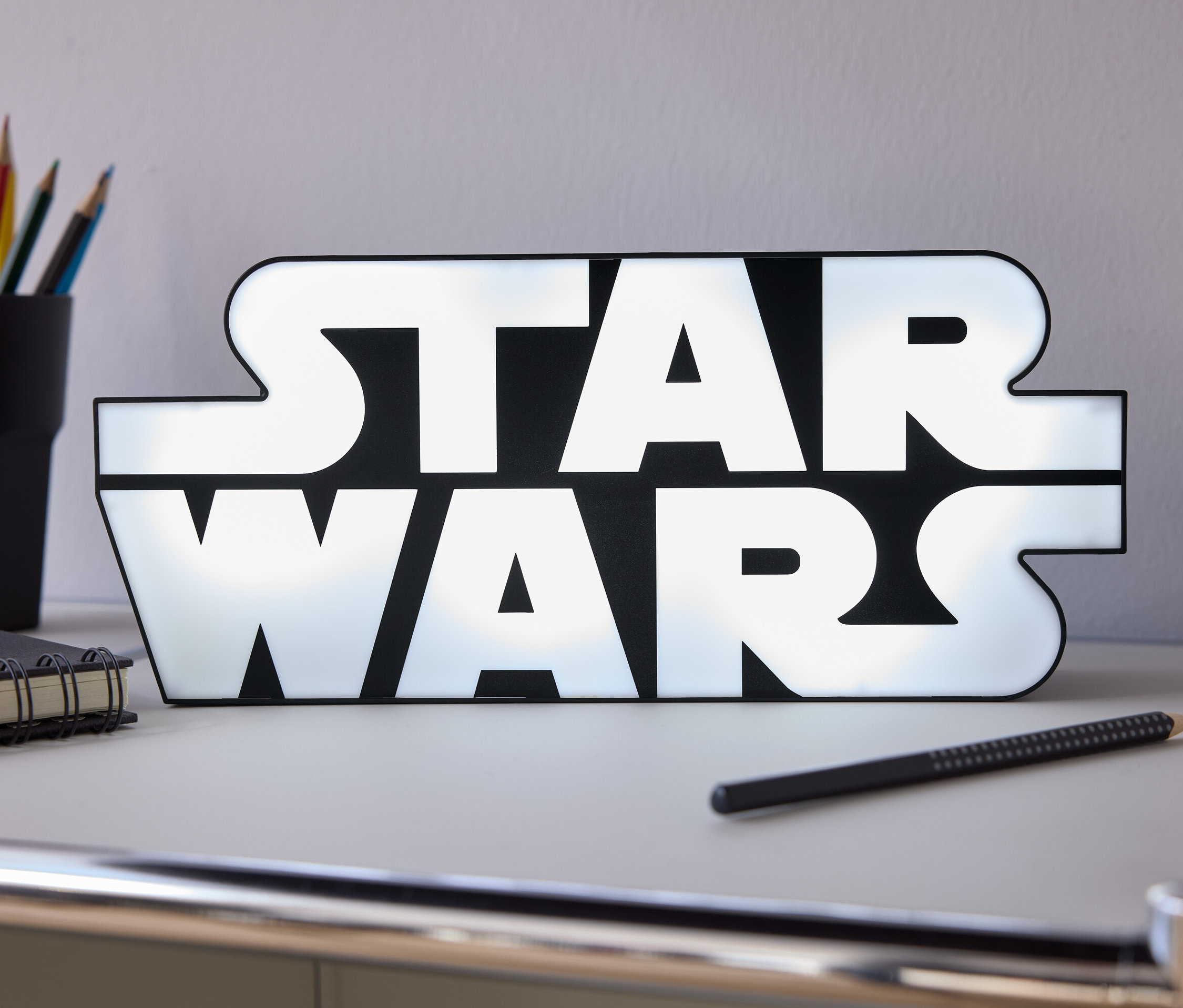 Sur une table, il y a une enseigne lumineuse Star Wars, un crayon, un carnet et un pot avec des crayons de couleur.