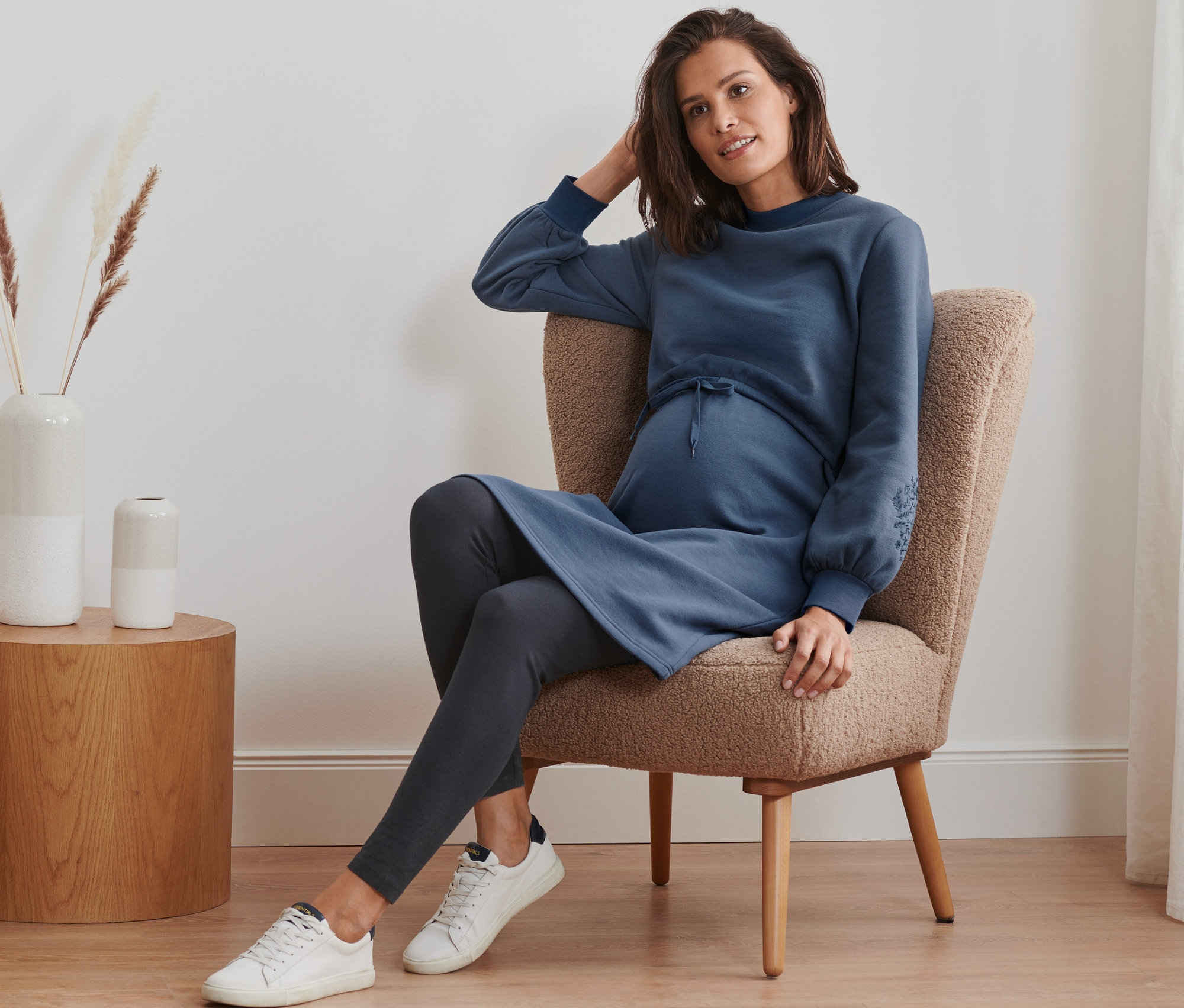 Une femme enceinte est assise dans un fauteuil et porte une robe molletonnée de grossesse et d'allaitement, des leggings et des baskets.