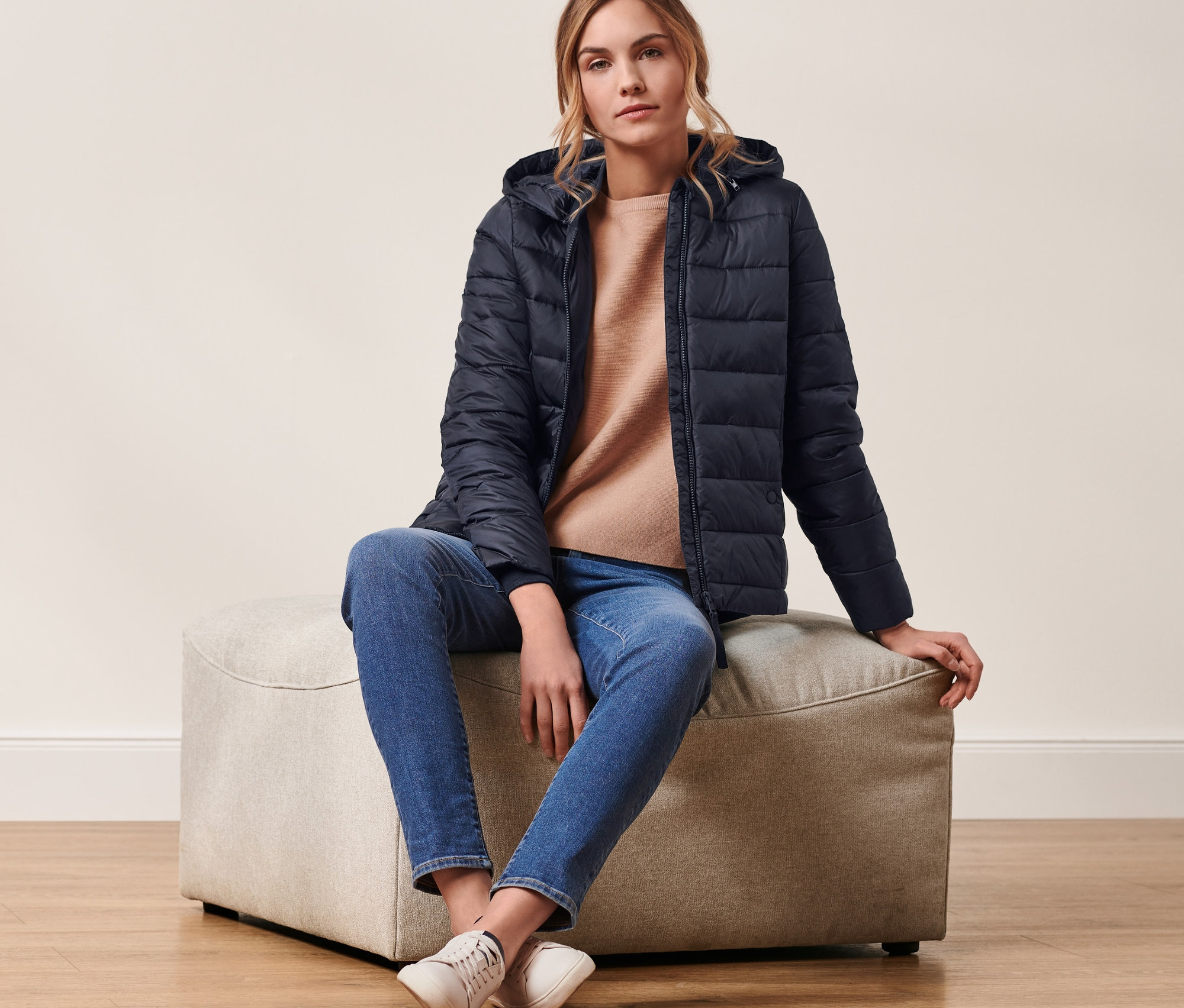 Une femme assise sur un tabouret porte une veste matelassée à capuche bleu foncé.