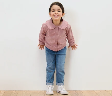Une fillette souriante se tient devant un mur blanc, vêtue d'un jean et d'une veste en sweat avec volant pour enfant.