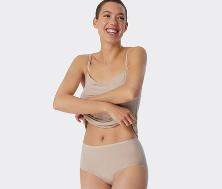 Une femme souriante tient l'ourlet de son débardeur. Elle porte 2 midi slips SCHIESSER avec modal, couleur sable, à rayures.