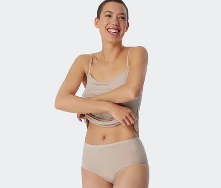 Femme souriante aux cheveux courts remontant son débardeur couleur sable, portant un slip midi SCHIESSER en modal couleur sable.