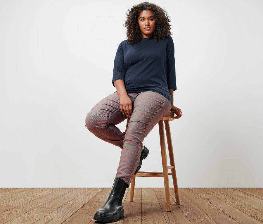 Une femme est assise sur un tabouret et porte un t-shirt manches 3/4 bleu foncé et un pantalon stretch à carreaux.