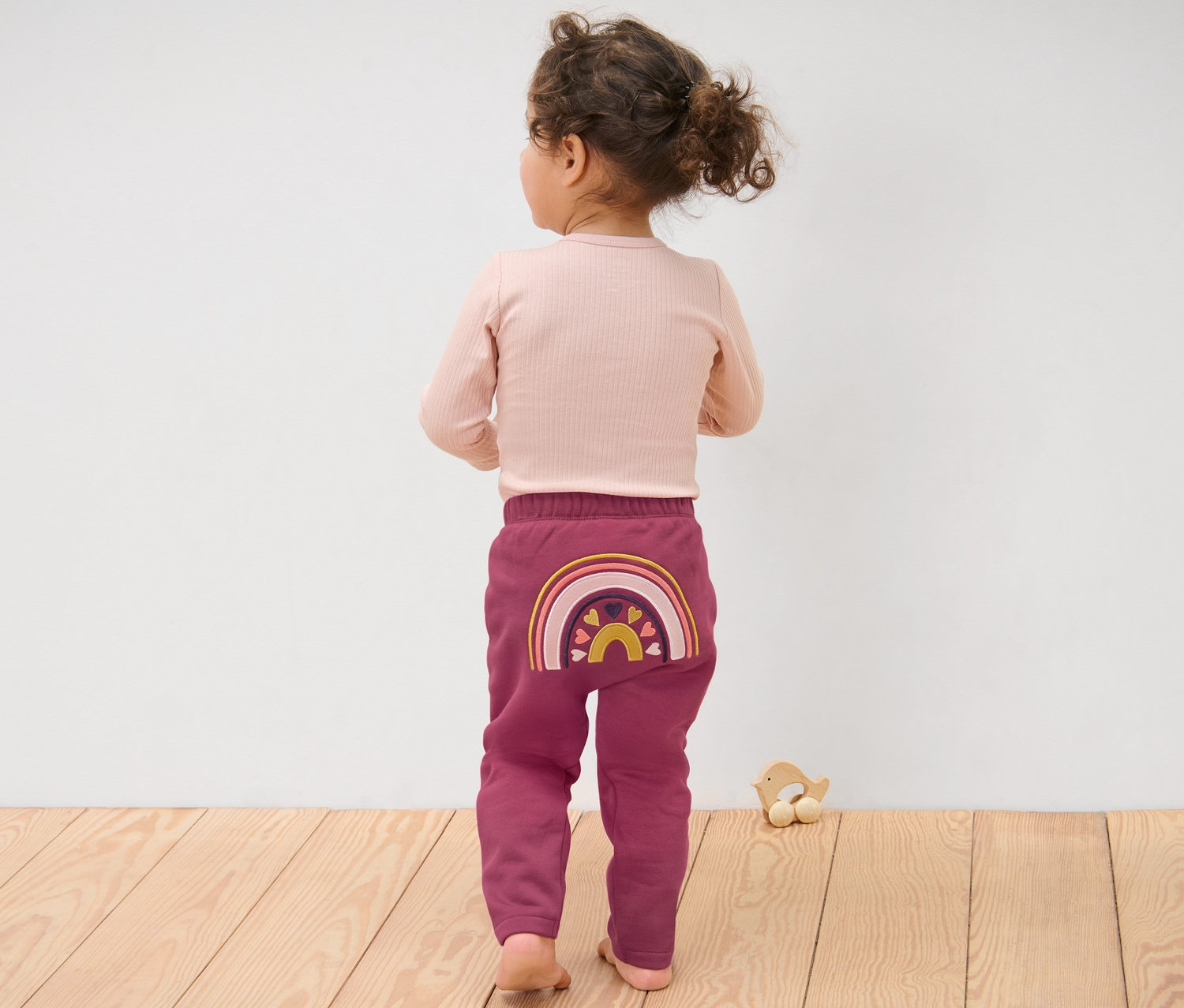 Fillette portant un pantalon en sweat pour bébé, arc-en-ciel, debout, dos à la caméra.