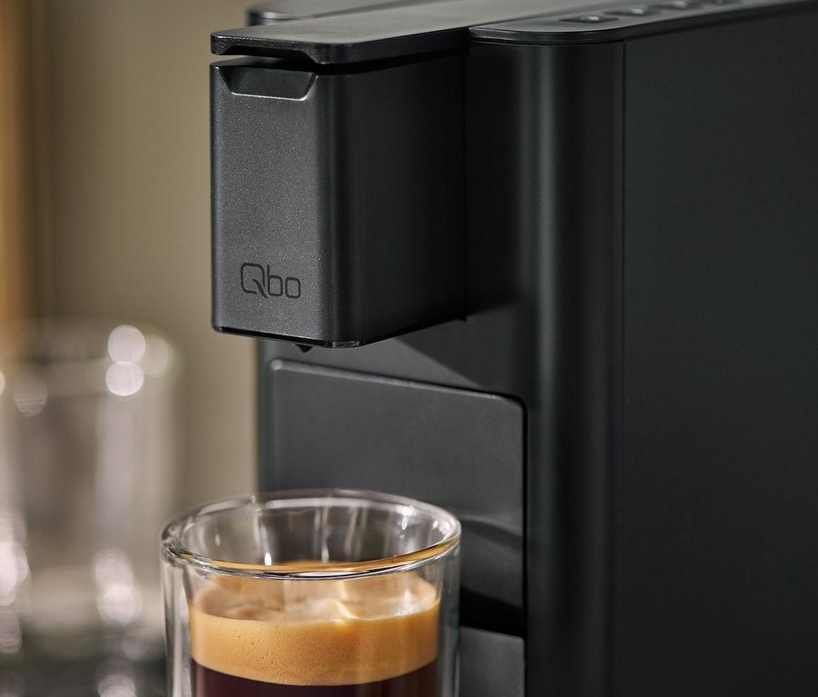 Gros plan d'une machine à café Qbo ICONIC Dark Chrome avec une tasse remplie de café.