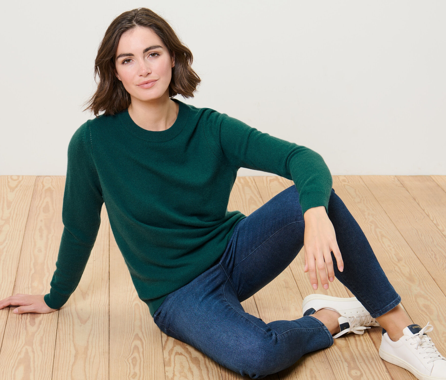 Une femme est assise par terre et porte un pull en maille fine vert avec du cachemire et un jean bleu.