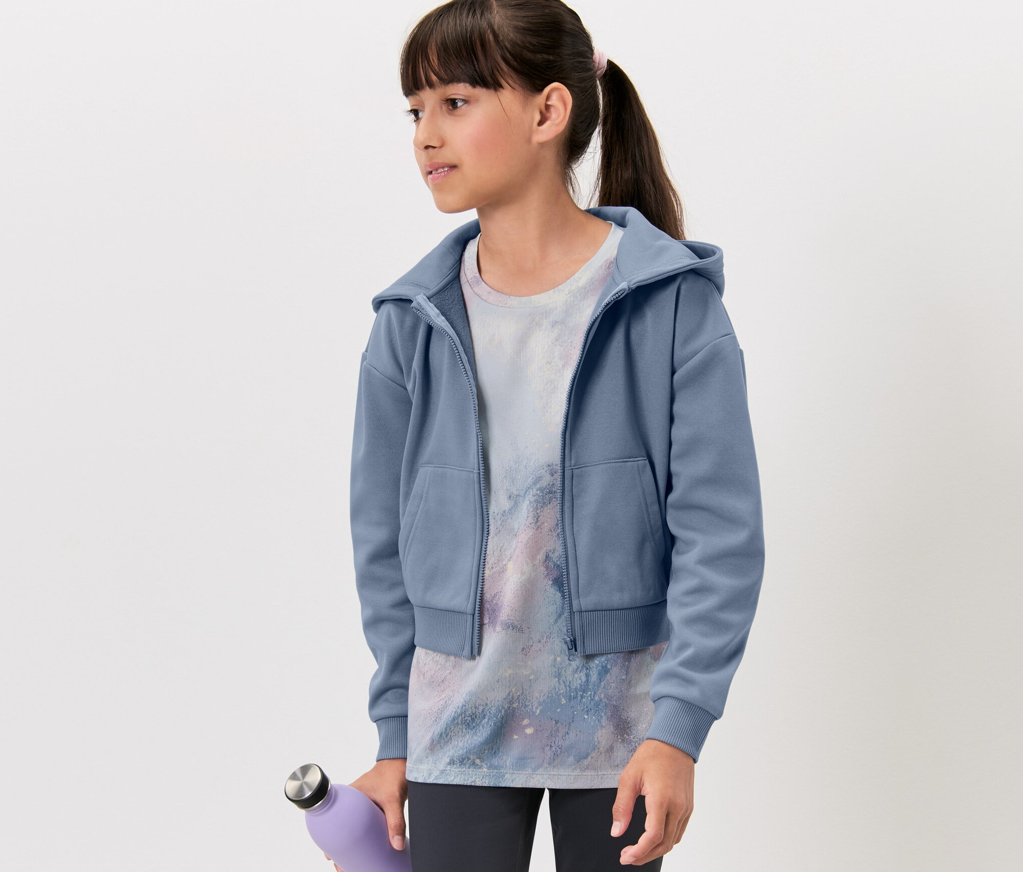 Une fille porte une veste de sport pour enfant, un t-shirt imprimé et tient une bouteille violette.