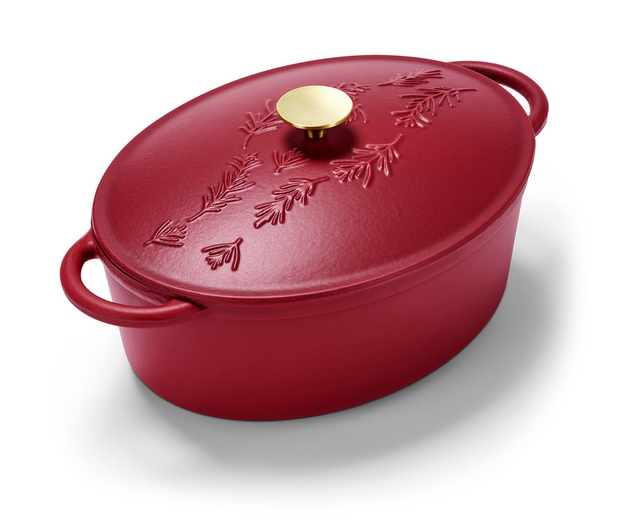 Une cocotte en fonte ovale rouge avec un motif de branches de sapin et une poignée dorée.
