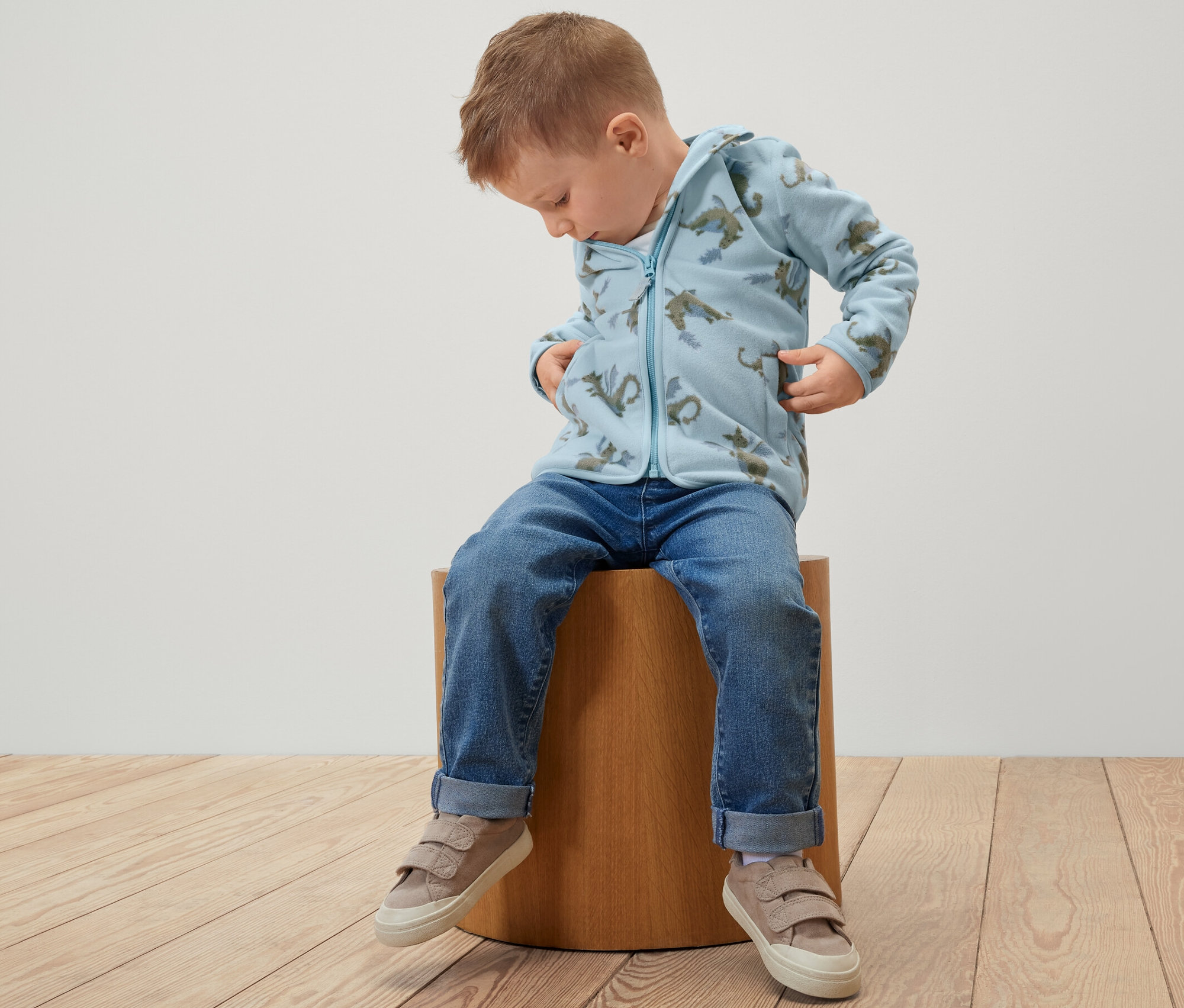 Un garçon est assis sur un tabouret et porte une veste polaire pour enfant avec un motif de dragon.