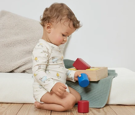 Un bambin bouclé est assis par terre et joue avec des blocs de couleur. Il porte 3 bodys pour bébé avec fonction évolutive, bleu.