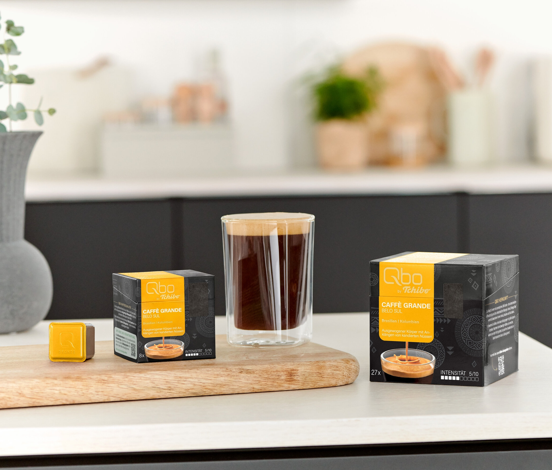 Un verre de café est posé à côté d'un paquet de Qbo Caffè Grande BELO SUL - 8 capsules et d'une capsule individuelle sur une planche en bois.