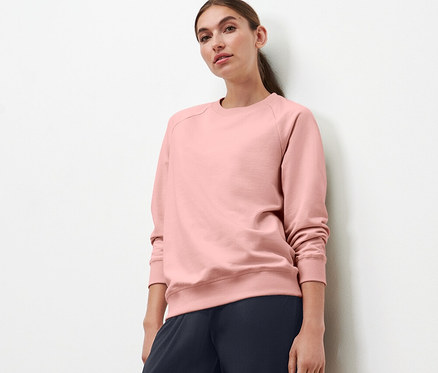 Une femme debout devant un mur blanc porte un sweatshirt de yoga rose et un pantalon bleu marine.