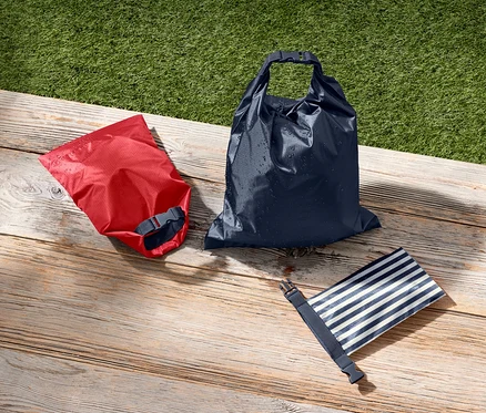 Un sac étanche rouge, un bleu marine et un bleu et blanc rayé sont posés sur une surface en bois à côté d'herbe verte.