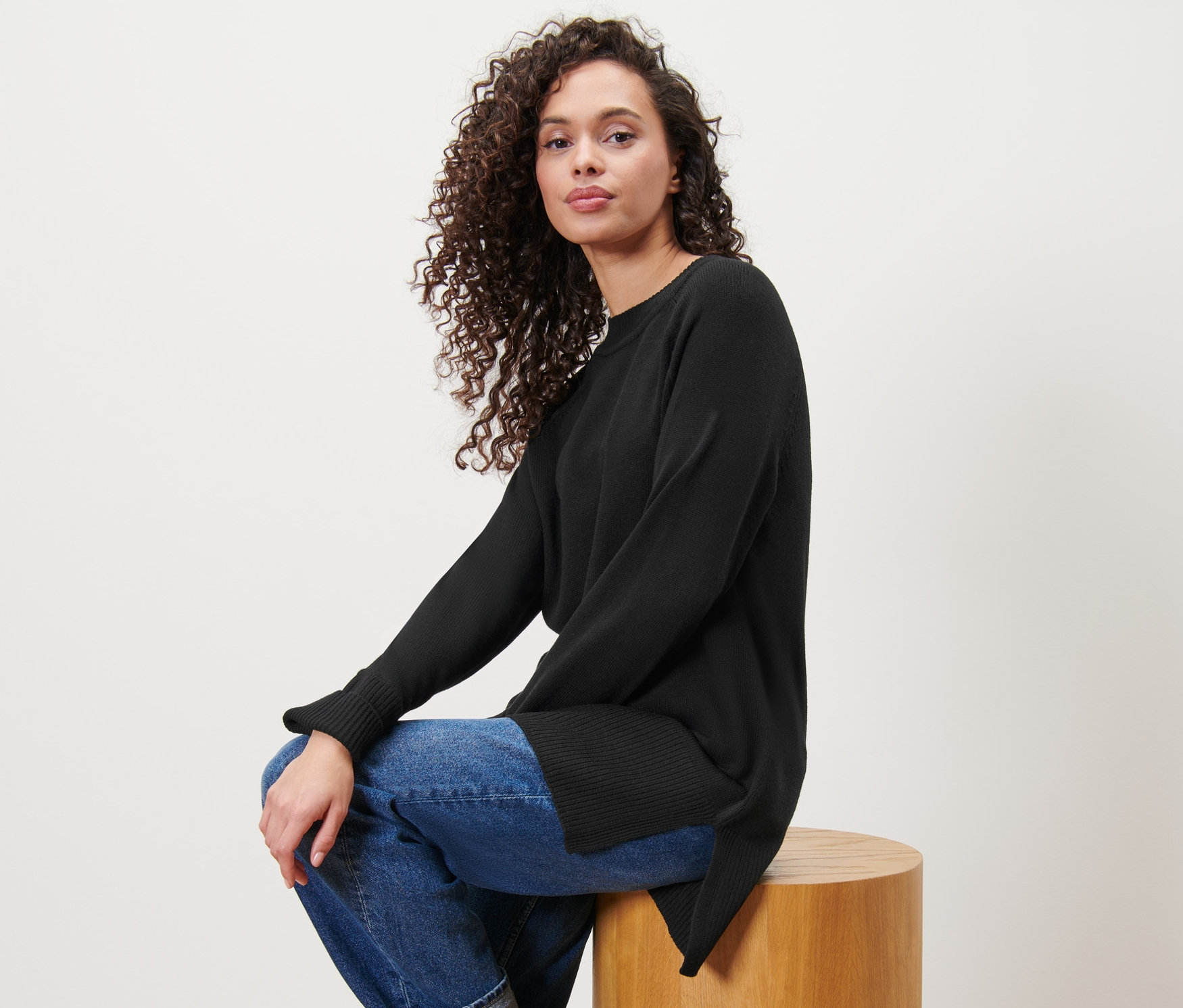 Femme assise sur un tabouret en bois portant un long pull en maille noir.