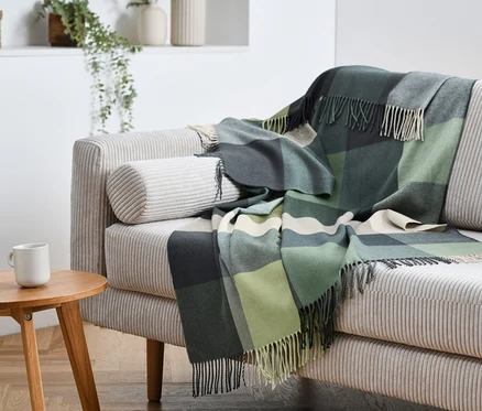 Un plaid vert à carreaux avec des franges est posé sur un canapé beige. Une tasse blanche est posée sur une table en bois.