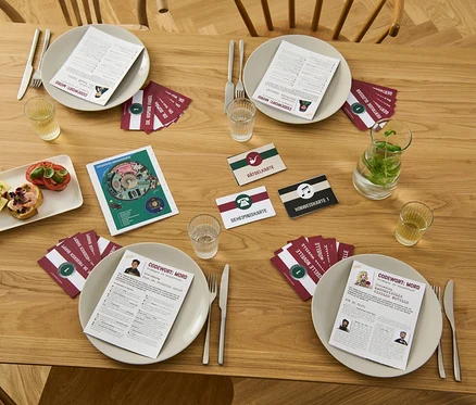 Sur une table en bois, des assiettes avec des cartes et des boissons pour le jeu d'enquête « Codewort: Mord – gefangen im Leuchtturm ».
