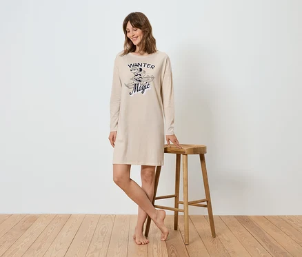 Une femme se tient debout et s'appuie sur un tabouret en bois. Elle porte une chemise de nuit beige à manches longues avec un imprimé.