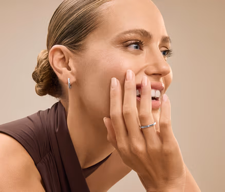 Portrait d'une femme aux cheveux relevés en chignon, souriant et touchant son visage avec une main portant une bague argentée.