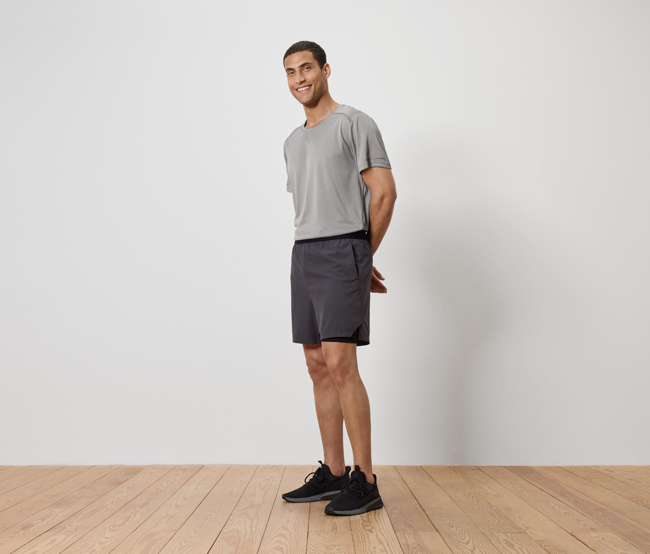 Un homme en t-shirt gris, short et baskets debout sur un plancher en bois devant un mur blanc.