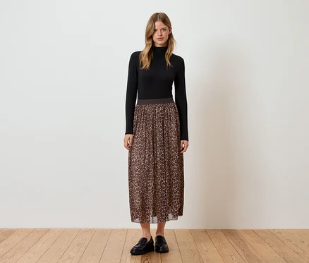 Une femme debout porte un col roulé noir, une jupe plissée marron à motif animalier et des chaussures noires.