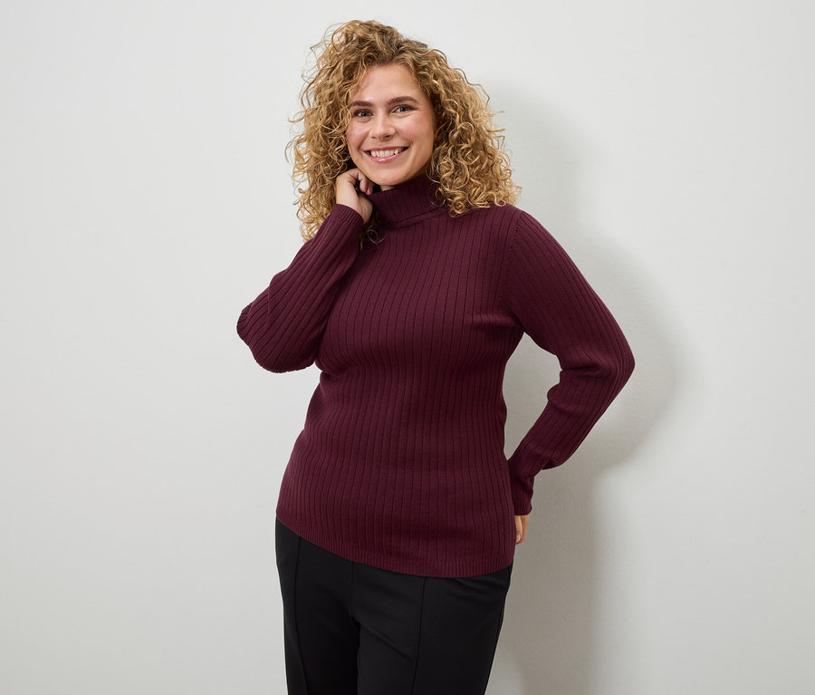 Une femme aux cheveux bouclés pose en pull à col roulé bordeaux et pantalon noir.